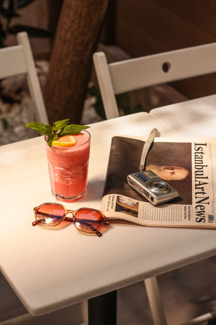A cool iced drink with lemon sits on a table with a newspaper and camera, evoking a relaxed summer vibe.