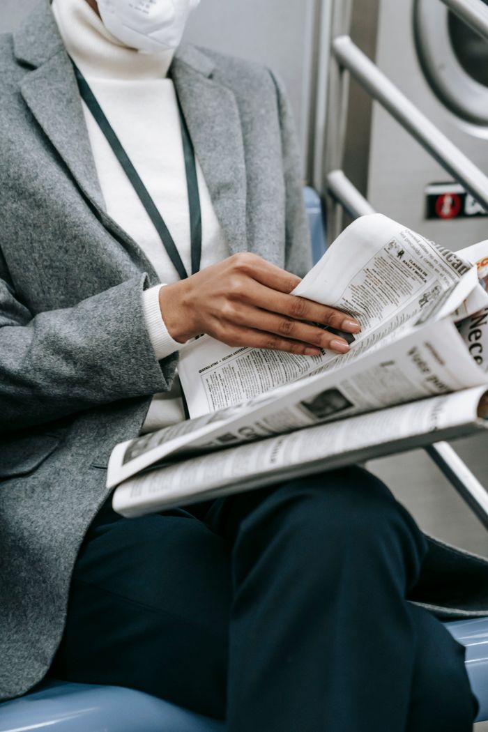 hero-img-01 Crop anonymous black female entrepreneur in elegant outfit and medical mask reading newspaper while riding train